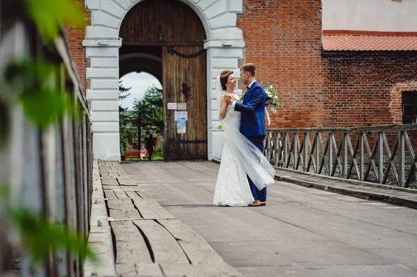 The fiance in a tuxedo and the bride in a white dress are dancing, against the background of an old brick building. Happy loving couple on wedding day outdoors