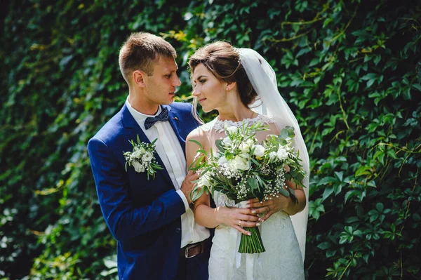 Bride and groom stand face to face against the backdrop of green nature. Happy loving couple on wedding day. Gorgeous wedding bouquet of white flowers in the hands of the bride. Soon kiss.