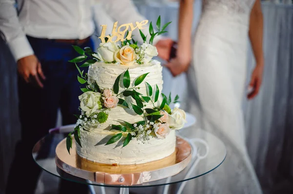 时尚的两层婚礼蛋糕 装饰着玫瑰 绿叶和其它花朵 矗立在一张玻璃桌子上 背景模糊 是新婚夫妇 很时髦的蛋糕 — 图库照片