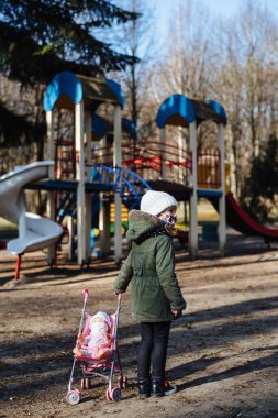 Kapalı oyun alanının arka planında koruyucu maskeli üzgün küçük kız. Tıbbi maskeli bir çocuk parkta yürüyor. COVID-19 'a karşı karantina önlemleri. Koronavirüs salgını.