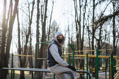 Çocuk parkında koruyucu maskeli ve eldivenli bir adam var. Gri kapüşonlu genç bir sporcu koronavirüs salgını sırasında parkta spor yapıyor. Ev aktivitelerinde kal. COVID-19.