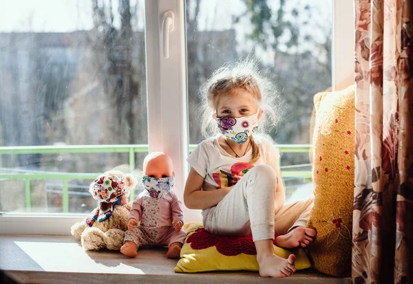 Koruyucu maske takan hasta bir çocuk pencerede oturur. Hasta enfeksiyonu önlemek için evde izole edilmiş. Coronavirus. Çocuğunuza covid-19, virüsler ve gribe karşı önleyici önlemler öğretmek.