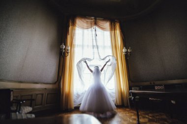 Gelin saray penceresinin yanında dururken perdelere dokunur. Perdeleri açan beyaz elbiseli güzel gelin. Arka görünüm.