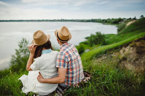 美しい若い男と女の愛の物語 恋人のカップルの男性と女性がお互いに川の銀行 バックビューに座って抱擁 帽子のスタイリッシュなカップル — ストック写真