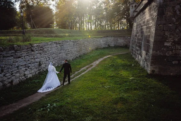 newlyweds walk through the courtyard of the medieval castle. The bridegroom in black tuxedo holds the bride in a white dress for her hand. Rear view Wide angle, top view. Wedding photo shoot.