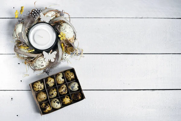 Pasen voorjaar decoratieve compositie, vervaardigde krans met kaars binnen en doos met kwarteleitjes. Close-up van portret op witte houten achtergrond met plaats voor tekst. — Stockfoto