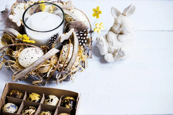 Easter spring decorative composition, crafted wreath with candle inside, white bunny and box with quail eggs. Close up portrait on white wooden background with place for text.