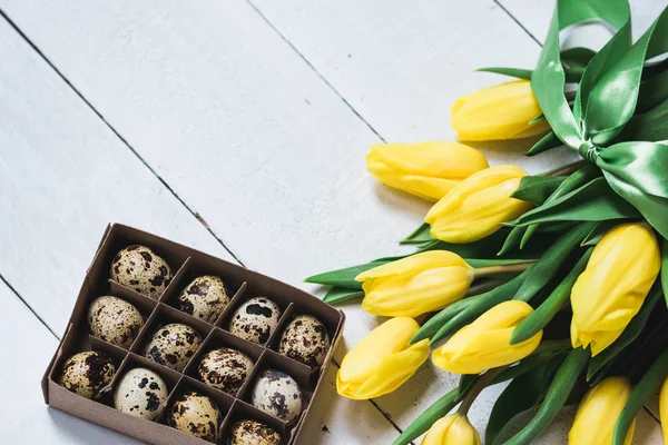 Pasqua primavera composizione decorativa. Bouquet di tulipani gialli legati da nastro verde e scatoletta con uova di quaglia. Ritratto ravvicinato su sfondo bianco in legno con spazio per il testo . — Foto Stock