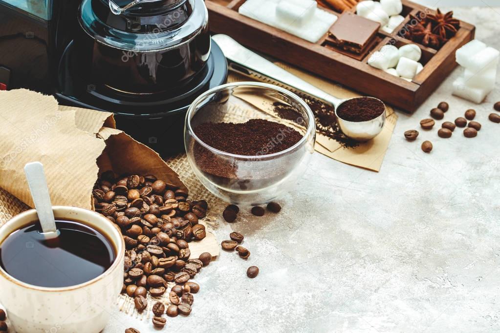 Variety things for prepare coffee. Roasted beans, ground coffee, scoop, electric coffee machine and assortment of sweets and spices to eat with on light background.