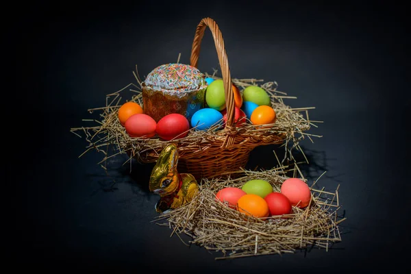 Easter colored eggs and Easter cake with straw in wicker basket on the black background