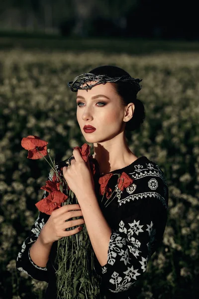 Portrait of young woman  with a  barbed wire wreath and a bouquet of poppies in the field