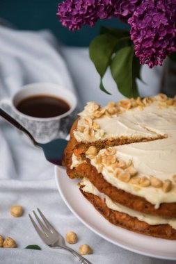 Fındıklı havuçlu kek, bir demet mor leylak ve beyaz masa örtüsünde bir fincan kahve havuçlu bisküvi ve fındıklı ekşi kremayla pişirilmiş turta.