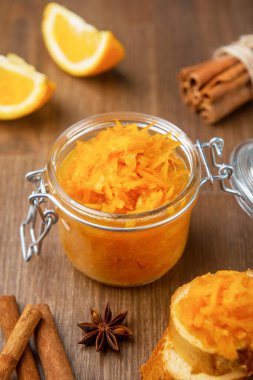 Tahta arka planda Fransız ekmeği, tarçın ve yıldız anason parçalarıyla dolu bir kavanoz turuncu konfeksiyon, ahşap masada birkaç parça ekmek bulunan ev yapımı portakal reçeli.