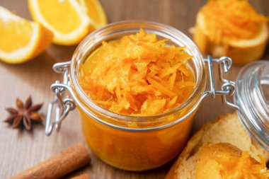 Tahta arka planda Fransız ekmeği, tarçın ve yıldız anason parçalarıyla dolu bir kavanoz turuncu konfeksiyon, ahşap masada birkaç parça ekmek bulunan ev yapımı portakal reçeli.