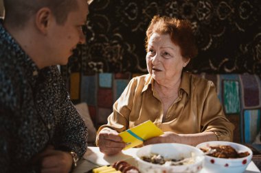 Torun, büyükannesiyle masaya oturuyor ve yemek yiyor. Torunu büyükannesini ziyaret ediyor. Ev yemeklerini yiyor.
