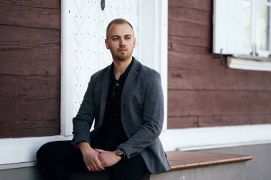 Sakallı genç adam binanın yanındaki merdivenlerde oturuyor ve yan tarafa bakıyor.