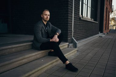 Ceketli, siyah binanın yanındaki merdivenlerde oturan yakışıklı, ciddi bir iş adamı.