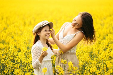 İki kız kardeş kolza tohumu tarlasında şapkayı dener, sarı çiçeklerde fotoşizofreni