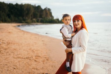 Kızıl saçlı genç bir kadın oğlunu deniz kenarında tutuyor.