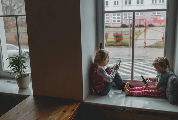 two sisters play a mobile phone on the sill of a large window. girls play with gadgets and have cute faces. the concept of a happy childhood. in the apartment,children play for a long time on the pho