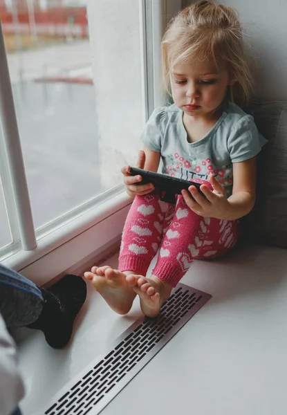 Küçük sarışın bir kız büyük bir pencerede evde oturuyor ve cep telefonuyla uzun süre oynuyor. Şu alete bak. modern çocukluk, tatlı yüz
