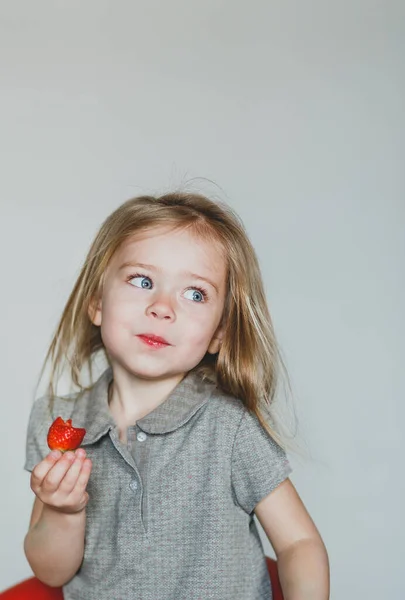 Задумчивая Маленькая Девочка Огромными Голубыми Глазами Сером Фоне Ест Клубнику — стоковое фото