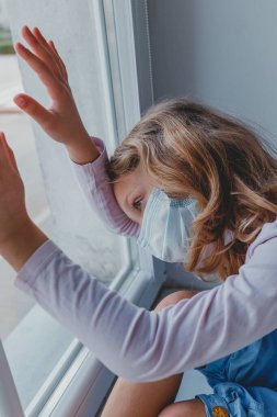 Üzgün bir Avrupalı kız elinde koronovirüs maskesiyle pencereden dışarı bakar.