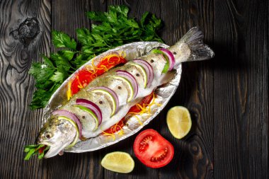 Çiğ balık alabalığı, limon ve folyolu bir tabakta domatesle pişirmek için pişirilir. Koyu bir ahşap arka planda.