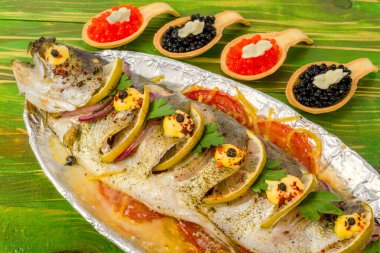 Balık alabalığı, limon, domates ve yeşillikle folyoda ızgara. Sırada kırmızı havyarlı turtalar var. Kaşık şeklinde. Yeşil ahşap arka planda.