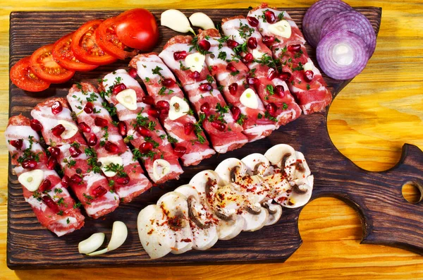 Damarlı domuz pastırması şeritlerine sarılmış kıymalı, sulu, çiğ domuz eti ruloları hazırlıyorum. Et yemeği. Bitki ve nar tohumları serpiştirilmiş. Yakın domates, sarımsak ve doğranmış mantar. Tahta kesme tahtasının üzerinde. Yakın plan..