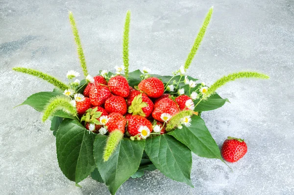 Yapraklarla süslenmiş sepetin içinde olgun çilekler ve gri arka planda tarla çiçekleri. Cömert bir yaz çilek hasadı konsepti. Yakın plan..