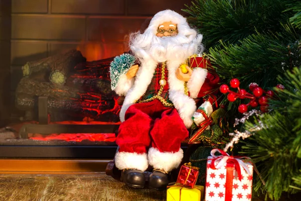 Şöminenin yanında ve Noel ağacında oyuncak bir Noel Baba oturuyor. Noel ağacının altında Noel hediyeleri yatıyor. Arka plan bulanık. Seçici odaklanma. Yakın plan..