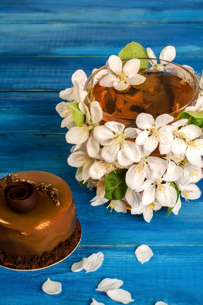 Elma çiçekleri ve güzel bir çikolatalı pastayla süslenmiş bir fincan çiçek çayı mavi bir ahşap masada yatıyor. Bahar içeceği kavramı. Dikey. Yakın plan..