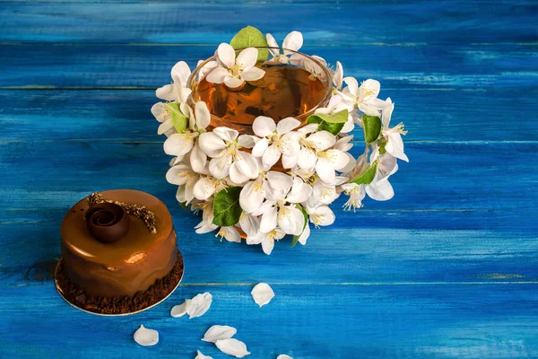 Elma çiçekleri ve güzel bir çikolatalı pastayla süslenmiş bir fincan çiçek çayı mavi bir ahşap masada yatıyor. Bahar içeceği kavramı. Yakın plan..