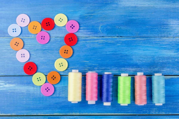 Çok renkli düğmeler ve iplikler, kalp şeklinde dizilmiş, aynı zamanda mavi ahşap arka planda iğneler. Üst Manzara. Düz görüntü. Yakın plan.. 
