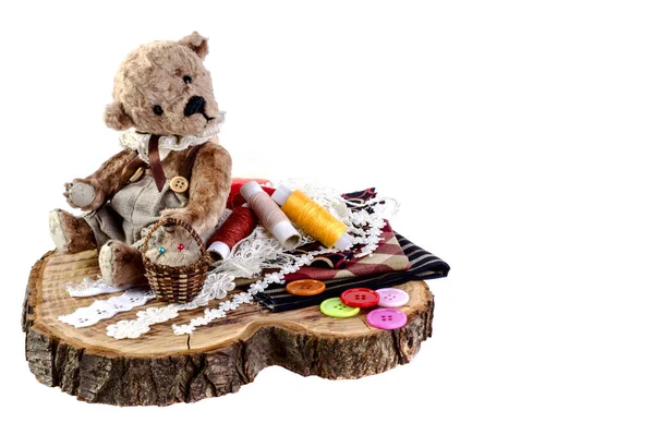 Şirin ayıcık kütüğün üzerinde oturuyor. Yanında kumaş, düğme ve iplik parçaları var..