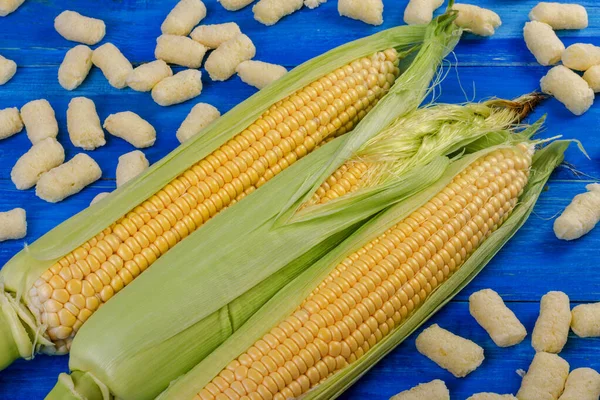 Three cobs of ripe sweet corn lie on a blue wooden background. Corn sticks are scattered around.