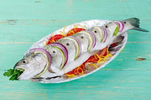 Çiğ balık alabalığı, limon ve folyolu bir tabakta domatesle pişirmek için pişirilir. Mavi ahşap bir arka planda.