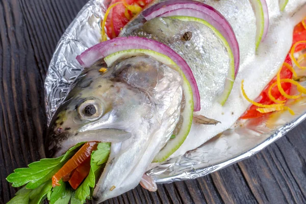Çiğ balık alabalığı, limon ve folyolu bir tabakta domatesle pişirmek için pişirilir. Koyu bir ahşap arka planda.