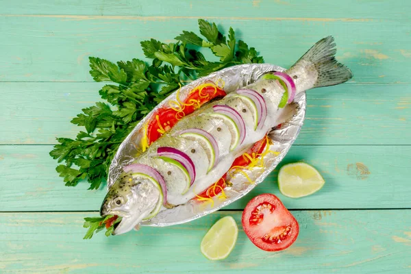 Çiğ balık alabalığı, limon ve folyolu bir tabakta domatesle pişirmek için pişirilir. Mavi ahşap bir arka planda.