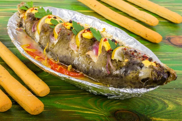 Balık alabalığı, limon, domates ve yeşillikle folyoda ızgara. Ekmek çubuklarının yanında. Yeşil ahşap arka planda. 