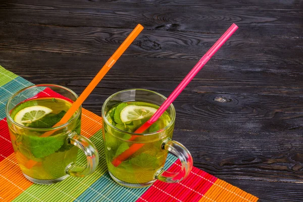 Nane ve limonlu soğuk çay kokteyli. Koyu, ahşap arka planda iki cam bardakta. Kırmızı ve turuncu tüp ile çok renkli bir kamış peçetede..