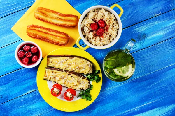 Sağlıklı beslenme kavramı. Sarı kasede ahududu reçeli yulaf ezmesi, sarı tabakta fırında patlıcan, iki kek ve mavi ahşap arka planda şeffaf bir fincanda naneli çay. Üst Manzara. Yakın plan..