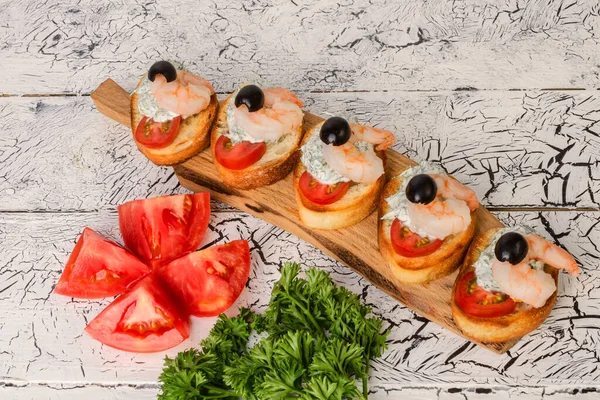 Tahta kesme tahtasının üzerinde karidesli ve domatesli lezzetli kanepeler. Ve yakınlarda bir sürü maydanoz var. Çizik beyaz ahşap arka planda.