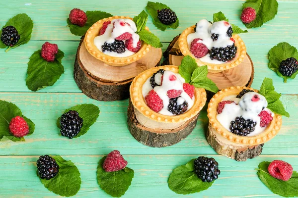 Böğürtlenli ve kremalı tart, dekoratif kütüklerin üzerine nane yaprağı. Etrafında da yapraklar ve böğürtlenler var. Mavi ahşap bir arka planda.