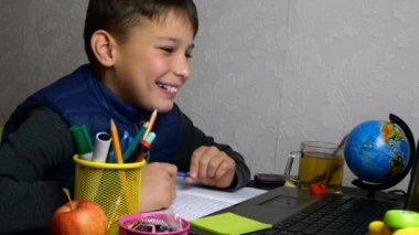 Uzaktan öğrenme, E-öğrenme. Öğrenci internetten eğitim alıyor ve karantina sırasında not defteriyle okul ödevi yapıyor. Masaüstünde okul malzemeleriyle oturuyor. Yakın plan portre.