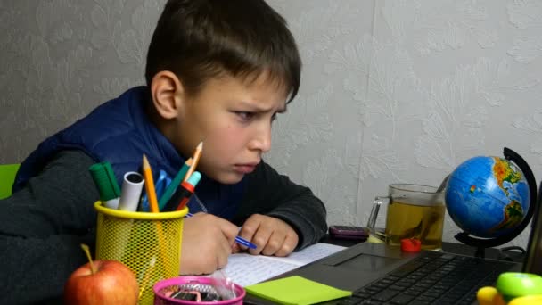 Afstandsonderwijs. Grappige schooljongen krijgt online onderwijs en doet huiswerk op school tijdens quarantaine met notebook. Hij is aan het griezelen en rotzooien. Close-up portret. — Stockvideo