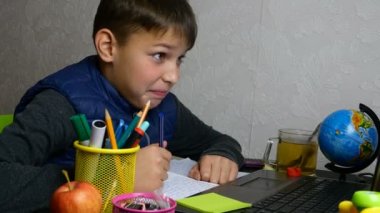 Mesafe öğrenme. Komik okul çocuğu internetten eğitim alıyor ve karantina sırasında not defteriyle okul ödevi yapıyor. Suratını ekşitiyor ve dalga geçiyor. Yakın plan portre.