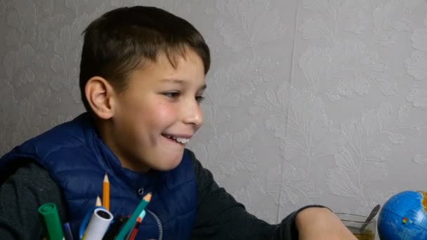 Afstandsonderwijs. Grappige schooljongen krijgt online onderwijs en doet huiswerk op school tijdens quarantaine met notebook. Hij is aan het griezelen en rotzooien. Close-up portret. — Stockvideo