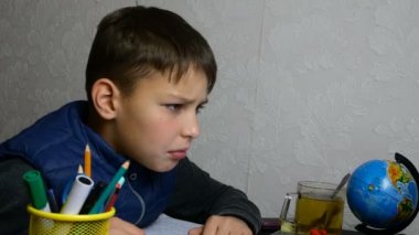 Uzaktan öğrenme, E-öğrenme. Ciddi okul çocuğu internetten eğitim alıyor ve karantina sırasında okul ödevi yapıyor. Masaüstünde okul malzemeleriyle oturuyor. Yakın plan portre.